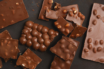 The chunks of broken different chocolate bars with nuts on the black stone surface with space for text. Background with chocolate. Sweet food photo concept.