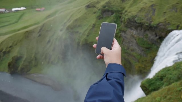 Man Taking Pictures of Famous Skogafoss Waterfall, Iceland With Smartphone, Close Up Slow Motion Pull Back. Social Media Infulencer Concept