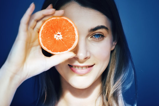 Beautiful Woman With Perfect Skin Hiding Eye Behind Orange Slice On Blue Background.