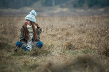 Cute white caucasian teen girl in a blue hat sitting in the middle of the field in cold autumn weather. Lifestyle background.
