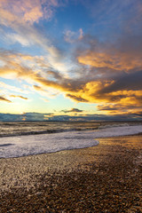 A view of a stony beach at sunset with crashing waves and ecume under a majestic yellow cloudy...