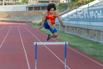 Obraz premium Latin fit young man with afro style hair in orange tank top at the running track jumping the hurdless fence on a sunny day.