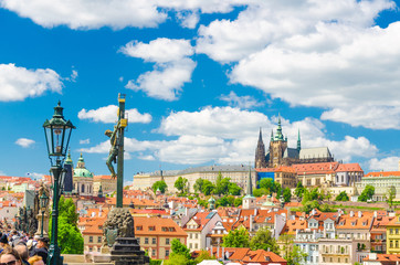Obraz premium Crucifix and Calvary statue on Charles Bridge Karluv Most over Vltava river with Prague Castle, St. Vitus Cathedral in Hradcany district, blue sky white clouds background, Bohemia, Czech Republic