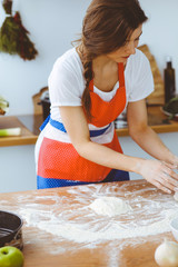Young brunette woman cooking pizza or handmade pasta in the kitchen. Housewife preparing dough on wooden table. Dieting, food and health concept
