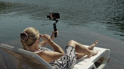 Woman shooting on handheld film gimbal stabilization for smartphone. Girl rest, relax, lie sunbed on pier in sunglasses, make selfie. Lady blogger broadcast video blogging vlogging. Take photo video. - obrazy, fototapety, plakaty
