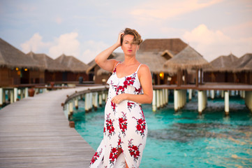 Pretty young woman wearing colorful dress posing near luxury water villas on tropical island resort