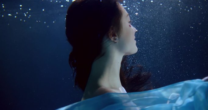portrait of a brunette with long flowing hair and a blue dress under water. she turns, puts her hand to her head, smiles. on the face there is a decoration of sequins. close up