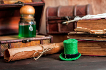 hat with green potion flask on wood table. Old books, bundle, box, chests, moss, shamrock. Saint...