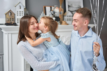 Fototapeta premium Loving family. Mom and dad hugging little daughter . Parents and baby child having fun near Christmas tree and white fireplace indoors. Merry Christmas and Happy New Year. Cheerful pretty people.