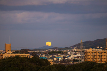 Fototapeta premium Full moon rises over distant mountain in small Japanese town at dusk