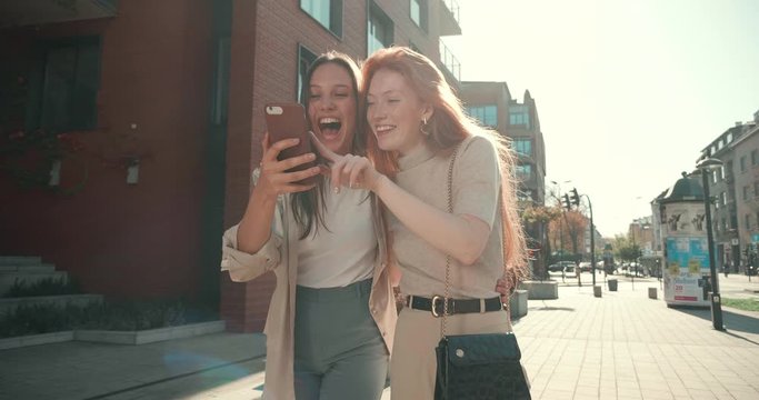 Couple of Female Young Friends walking in the Town while looking at Mobile Phone. Pretty women having Fun in the Center City. Smiling and Laughing while hugging each other. Happy Frienship.