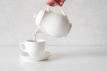 Pouring fresh tea from white teapot into white cup