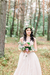Obraz na płótnie Canvas Beautiful bride with bouquet of flowers in white dress stands on forest background. Rustic style