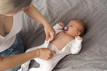 Mother with her newborn son laying on the bed in the rays of sunlight coming out of the window through the curtains. Mom dresses a newborn baby, buttoning up a jumpsuit. Home comfort.
