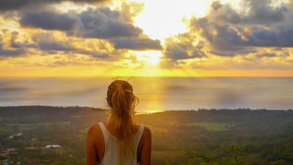 Inspiring scene of a woman standing at the viewpoint looking straight towards the pacific ocean during a stunning sunset making sky colourful and sun rays braking true the clouds. Feeling the freedom.