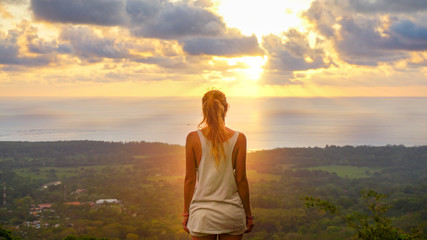 Inspiring scene of a woman standing at the viewpoint looking straight towards the pacific ocean during a stunning sunset making sky colourful and sun rays braking true the clouds. Feeling the freedom.