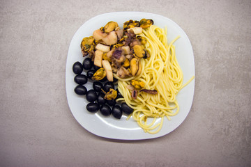 white plate on a gray kitchen table with spaghetti with black olives and seafood top view