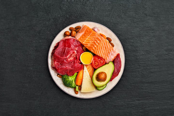 Plate with a ketogenic diet food. Beef, salmon, egg, broccoli, tomato, nuts, carrots, mushrooms, cucumber, dates. Diet food on a dark background with copy space. Top view. Flat lay