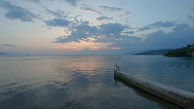 Rear view of little girl sitting on the edge of a dock and fishing with father Mediterranean Sea at sunset. Enjoying adventure in summer vacation