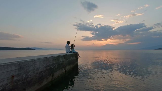 Rear view of little girl sitting on the edge of a dock and fishing with father Mediterranean Sea at sunset. Enjoying adventure in summer vacation