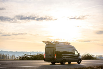 Dark compact cargo mimi van with ladders on the roof running on the road at sunset time