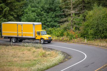 Fototapeta na wymiar Small compact yellow rig semi truck with long box trailer driving on the round highway entrance with green trees on the side