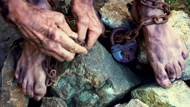 Man in captivity. A slave in iron chains is trying to free himself. A scene from the life of a slave. The symbol of slave labor. Hands and feet in chains. Breaking chains