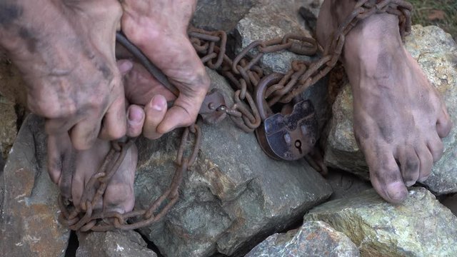 Man in captivity. A slave in iron chains is trying to free himself. A scene from the life of a slave. The symbol of slave labor. Hands and feet in chains. Breaking chains