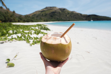 Woman's hand holding fresh coconut with bamboo straw on a beautiful white sandy beach in Fiji with turquoise water, green hills and plants, relaxing vacation destination