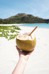 Woman's hand holding fresh coconut with bamboo straw on a beautiful white sandy beach in Fiji with turquoise water, green hills and plants, relaxing vacation destination