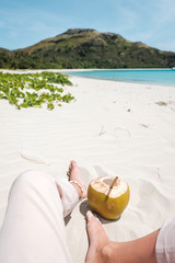 Fresh coconut with bamboo straw and girl's legs on a beautiful white sandy beach in Fiji with turquoise water, green hills and plants, relaxing vacation destination