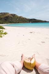 Fresh coconut with bamboo straw and girl's legs on a beautiful white sandy beach in Fiji with turquoise water, green hills and plants, relaxing vacation destination