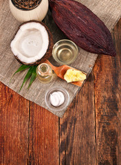Fototapeta na wymiar Half coconut with bottle and cup of coconut oil, cosmetic dispenser, cosmetic cream butter in scoop and cocoa pod on wooden background. Top view. Text space.