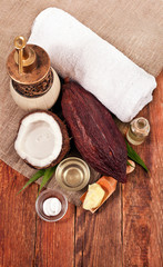 Half coconut with bottle and cup of coconut oil, cosmetic cream, butter in wooden scoop, towel and cosmetic dispenser on wooden background. Top view.