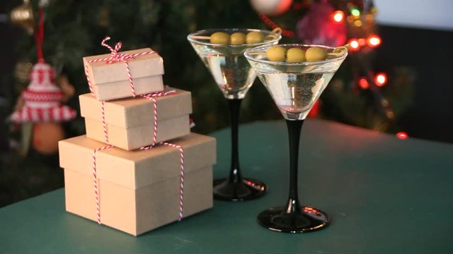 Two glasses of martini with olives and gift boxes on the green table. Light garland and Christmas tree with bokeh on the background. Woman hand put three various gift boxes