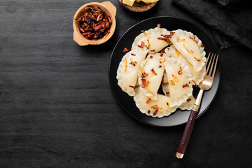 Homemade boiled dumplings filled with potato served with fried onion and bacon on black wooden background.
