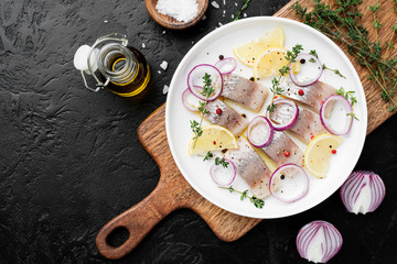 Salted herring with spices, lemon and red  onion rings on black stone background . Marinated sliced fish. top view