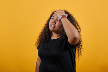 Serious afro-american young woman with overweight keeping hand on forehead, closed eyes over isolated orange background wearing fashion black shirt. People lifestyle concept.