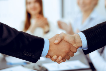 Business people shaking hands at meeting while theirs colleagues clapping and applauding. Group of unknown businessmen and women in modern white office. Success teamwork, partnership and handshake