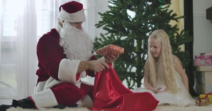 Portrait of beautiful blond Caucasian girl in white dress and crown sitting with Santa in front of New Year tree and taking off Christmas presents. Childhood, holidays season. Cinema 4k ProRes HQ.