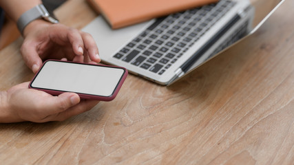 Cropped shot of young man using blank screen smartphone while working on his project with laptop
