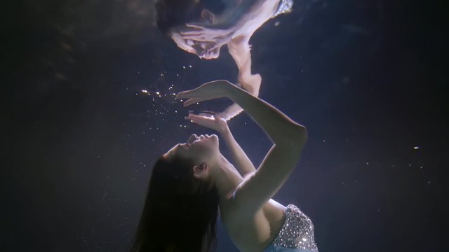 a girl with long dark hair and a sequined dress slowly approaches face up to her reflection on the surface of the water and passes through it