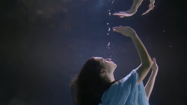 a brunette with long hair floats under the water. she reaches out her hand to the surface of the water and passes through it