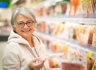 A smiling senior woman at the supermarket chooses which ham to buy. Many products on offer in the...