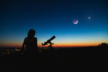 Astronomer with a telescope watching at the stars and Moon with blurred city lights in the...