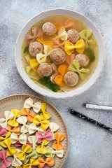 Plate of meatball soup with pasta and raw farfalle over beige stone background, above view, vertical shot