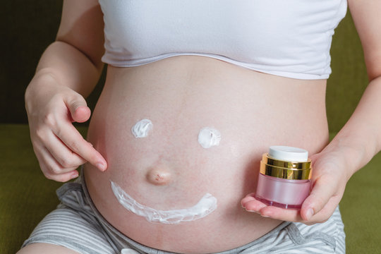 Pregnant Woman With Smile Is Using Moisturizing Cream On Her Tummy For Prevent Stretch Marks From Pregnancy While She Sits At Home ;Concept Of Pregnant Women Healthy And Health Care.