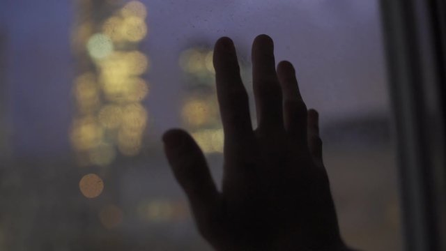 Hand entres frame and rests on glass window overlooking city night