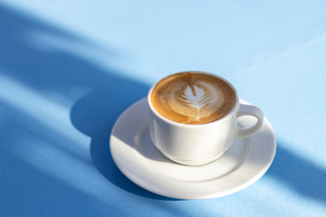 A cup of cappuccino on a light blue surface in sunlight.