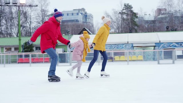 Side view tracking shot of happy family of mother, father and little daughter having fun ice skating together holding hands on outdoor skating rink in winter